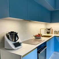 Błękitna kuchnia z białym blatem: Thermomix na pierwszym planie, złota armatura i owoce, eleganckie oświetlenie podszafkowe. Ujęcie z bliska, nowoczesny design.