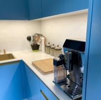 Błękitna kuchnia z frezowanymi frontami, złoty zlew, ekspres do kawy i drewniana deska na blacie. Nowoczesne oświetlenie podszafkowe.