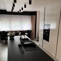 Nowoczesna kuchnia: czarny blat, białe szafki z połyskiem, oświetlenie punktowe, dekoracyjny wazon na wyspie, krzesła z pikowaniem w tle, minimalistyczny design.