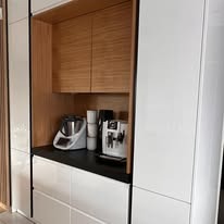 Biała zabudowa kuchenna z drewnianą wnęką, w której stoi ekspres do kawy, robot kuchenny i filiżanki. Minimalistyczny design, czarny blat, widok z boku.