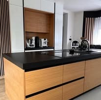 Nowoczesna kuchnia: drewniane fronty szafek dolnych, czarny blat, białe szafki górne, sprzęt AGD. Minimalistyczny design, pionowe panele dekoracyjne.