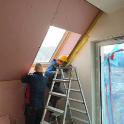 DAVI-BAU Dawid Orzechowski - Dwóch monterów na drabinie instaluje płytę gipsową wokół okna dachowego, widoczne ocieplenie i folia ochronna na oknie. Prace wykończeniowe na poddaszu.