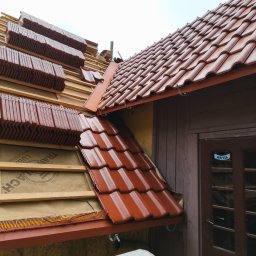 Davi-Bau - Częściowy widok dachu z czerwoną dachówką karpiówką w trakcie układania, widoczne deski i membrana. W tle fragment ściany budynku i drabina.