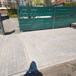 Davi-Bau - Jasnoszara kostka brukowa ułożona na podjeździe, z zielonym ogrodzeniem panelowym w tle. Widoczny cień osoby robiącej zdjęcie.