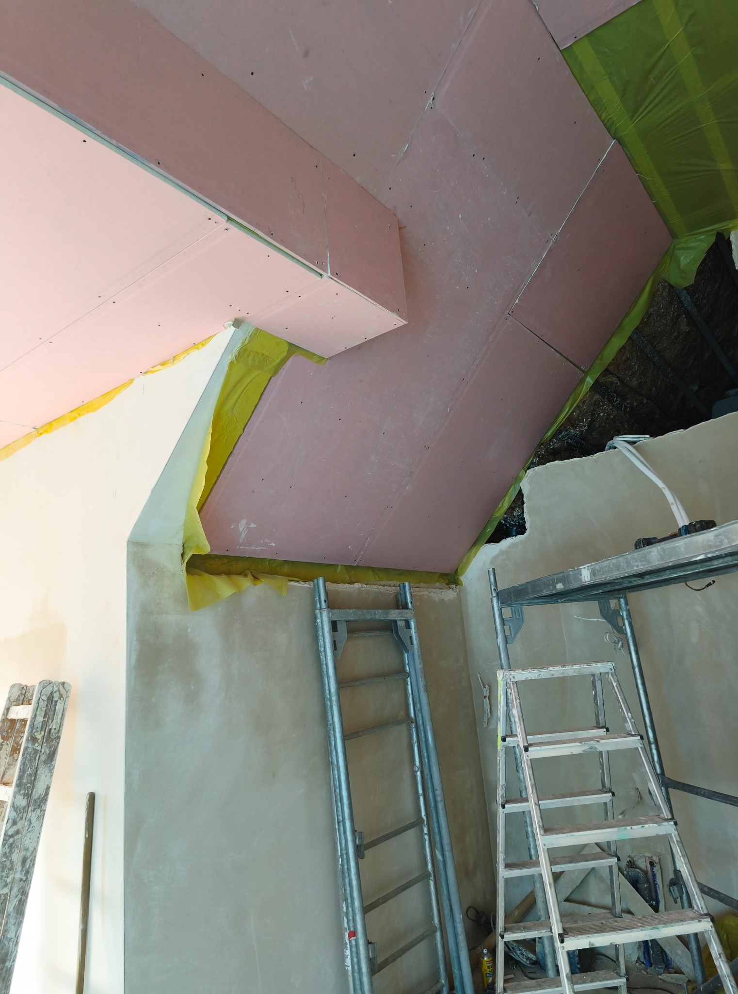 Skośny sufit poddasza w trakcie wykańczania płytami gipsowo-kartonowymi, widoczne rusztowania i folia ochronna. Ujęcie z dołu, podkreślające geometrię wnętrza.