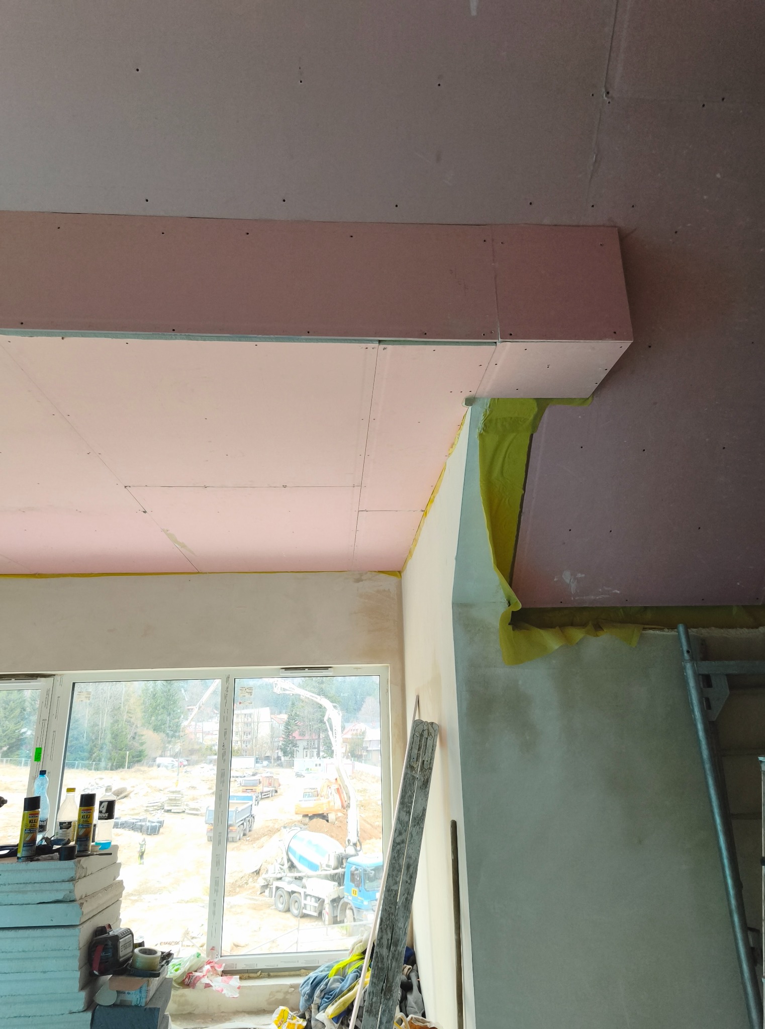 Montaż płyt gipsowo-kartonowych na suficie podwieszanym, widoczne profile i izolacja. W tle okno z widokiem na plac budowy z betoniarkami.