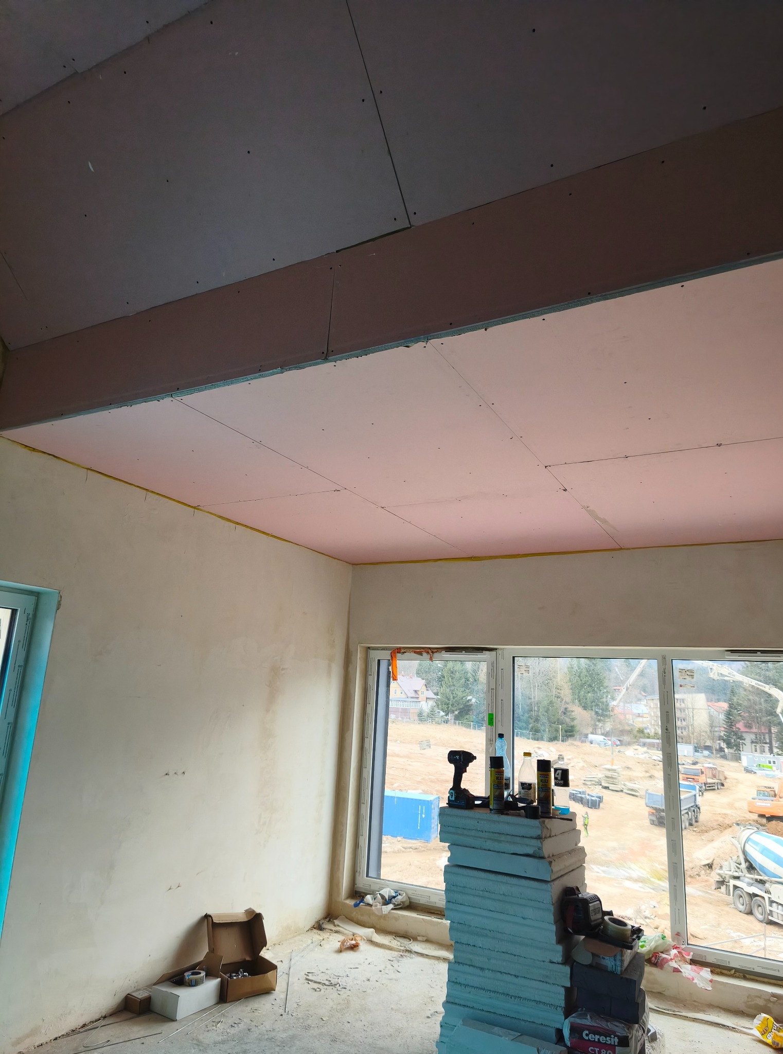 Montaż płyt gipsowo-kartonowych na suficie poddasza, widoczne narzędzia i materiały budowlane, okno z widokiem na plac budowy z betoniarką.