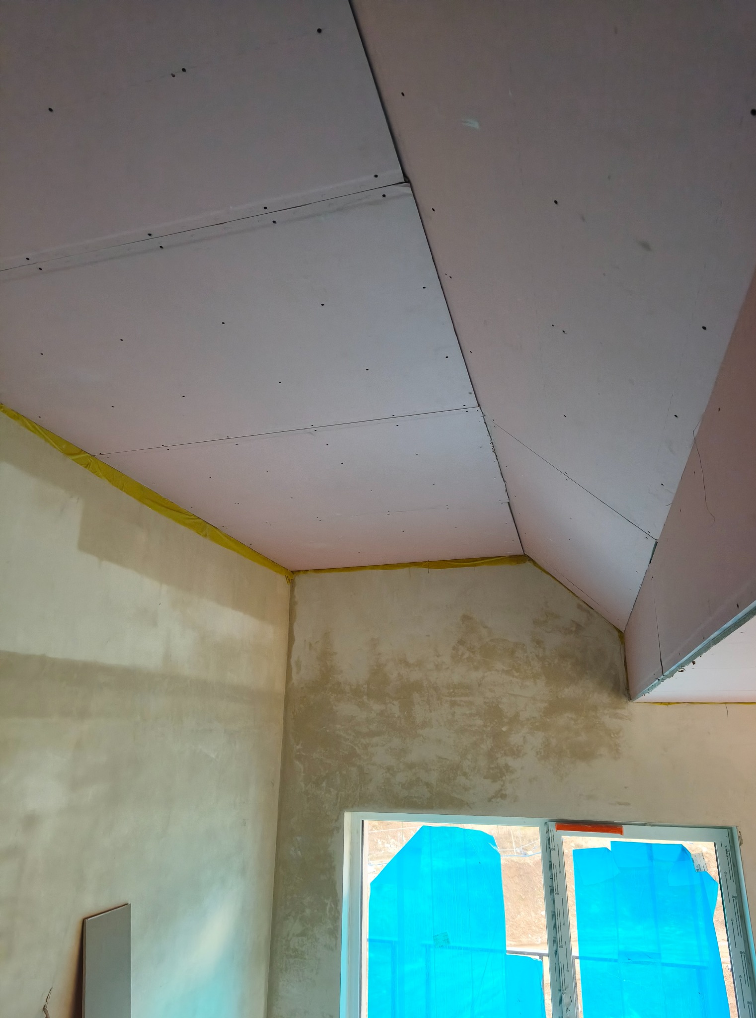 Skośny sufit poddasza z płyt gipsowo-kartonowych, mocowanych na stelażu. Ściany w stanie surowym, okno zabezpieczone folią. Żółta taśma na styku ściany i sufitu.