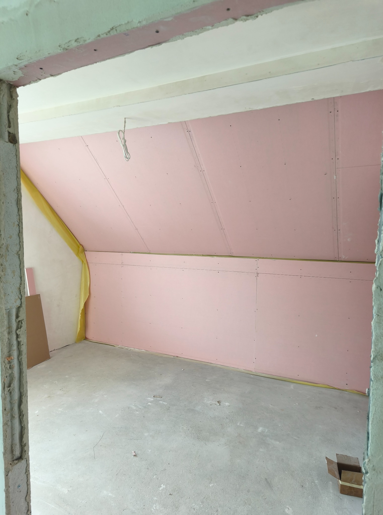 Poddasze w trakcie adaptacji: ściany i skosy wyłożone różowymi płytami gipsowo-kartonowymi, widoczne łączenia i mocowania. Surowy beton na podłodze, żółta taśma uszczelniająca.