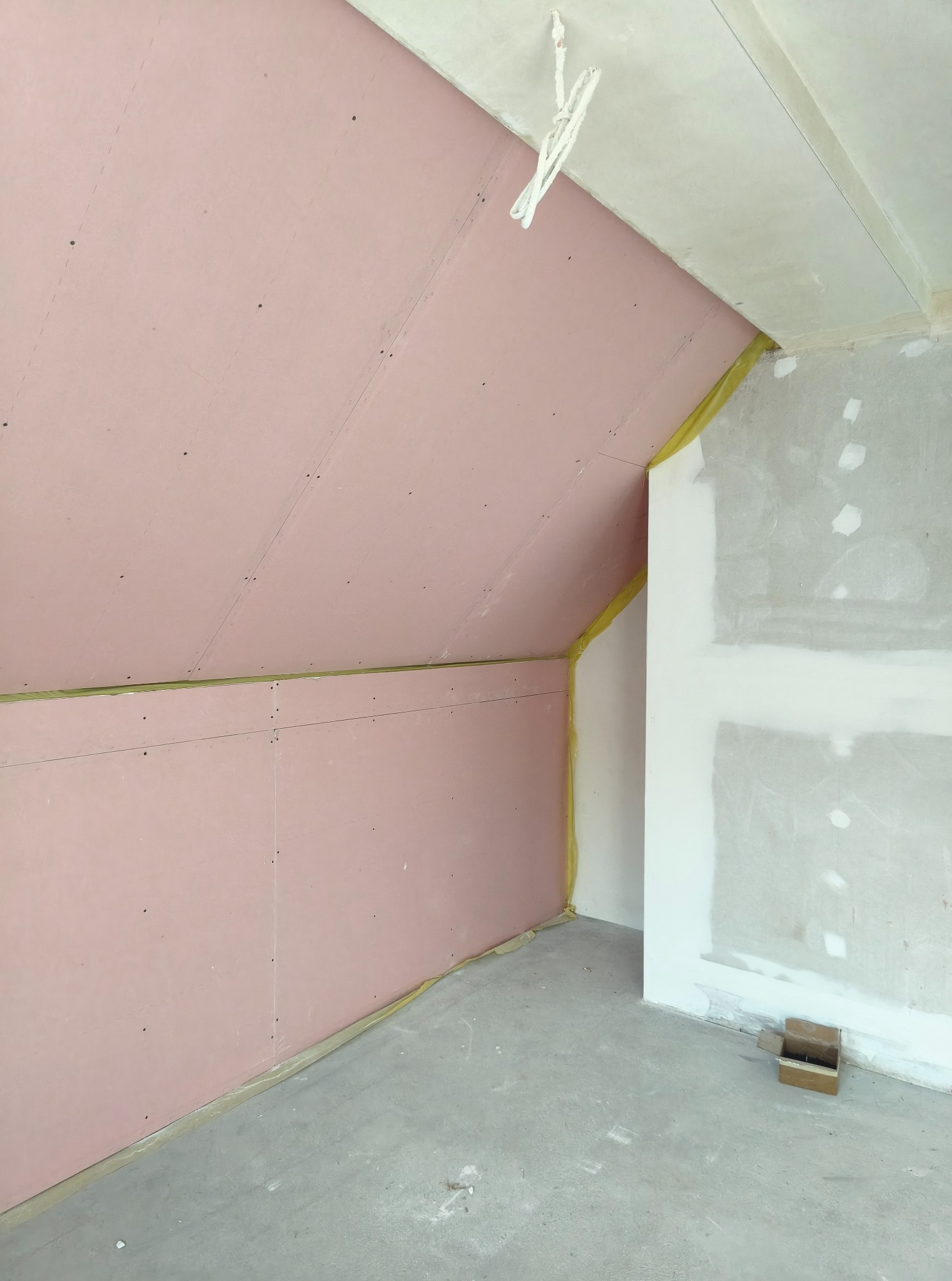 Poddasze w trakcie wykańczania: różowe płyty gipsowo-kartonowe na skosach, żółta wełna mineralna w szczelinach, surowy beton na podłodze, ściana częściowo zagruntowana.