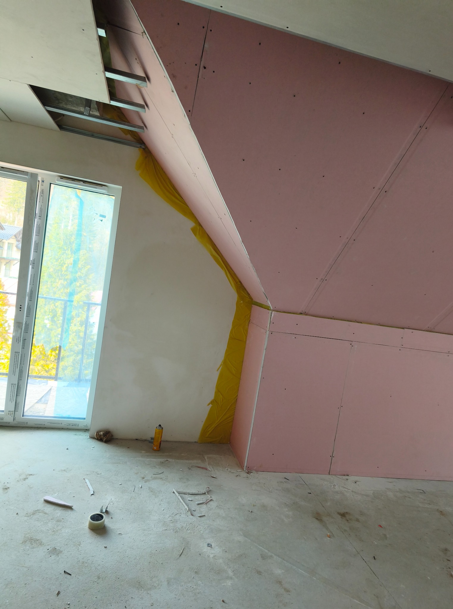 Poddasze w trakcie wykańczania: zamontowane płyty gipsowo-kartonowe na skosach, widoczne profile sufitowe i okno. Żółta wełna mineralna w szczelinach. Na podłodze resztki materiałów.
