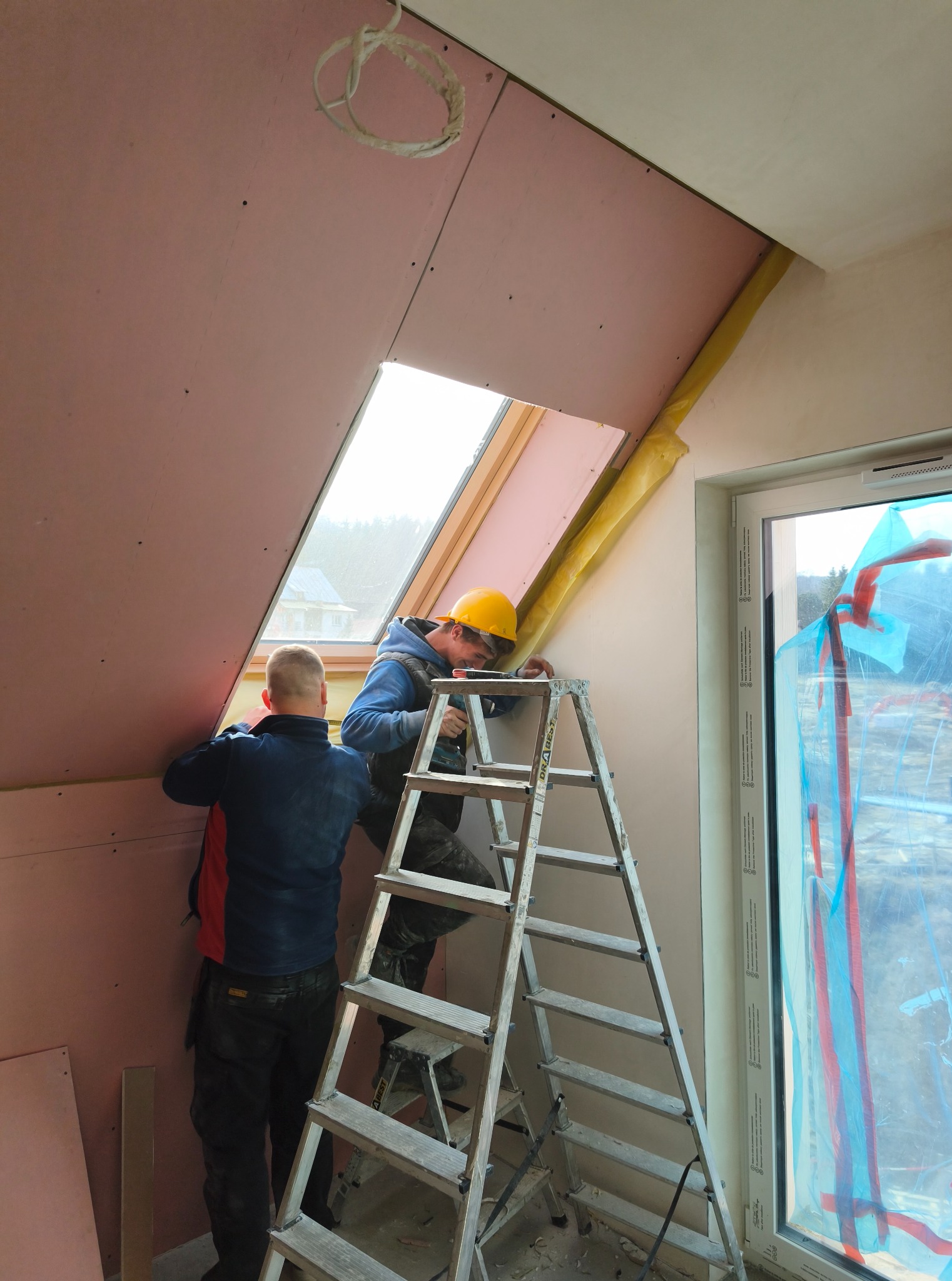 Dwóch monterów na drabinie instaluje płytę gipsową wokół okna dachowego, widoczne ocieplenie i folia ochronna na oknie. Prace wykończeniowe na poddaszu.