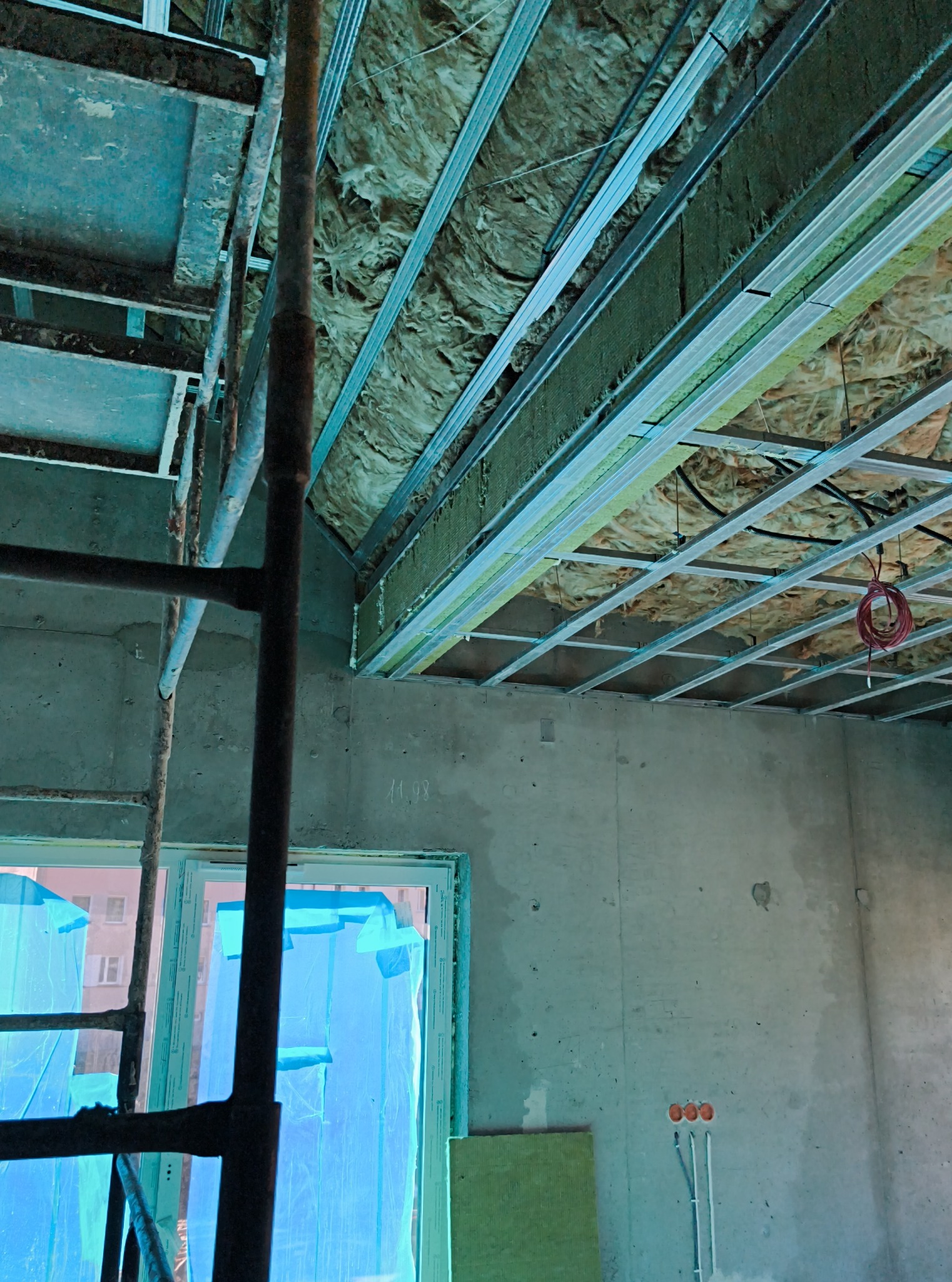 Montaż izolacji stropu z wełny mineralnej i metalowej konstrukcji. Widoczne rusztowanie, okno zabezpieczone folią i instalacja elektryczna. Prace remontowe w surowym wnętrzu.