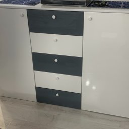 PS MEBLE Patryk Siwka - Nowoczesna komoda z szufladami i szafkami w kolorze białym i szarym, z okrągłymi uchwytami, zrealizowana w Gnieźnie. Na blacie widoczny odtwarzacz DVD i dekoracyjne serwetki.