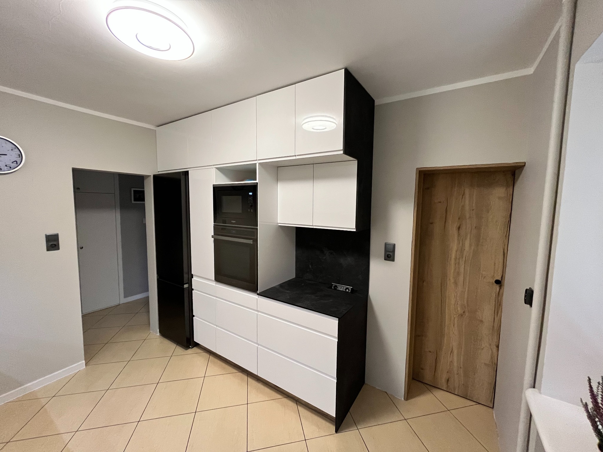 Nowoczesna kuchnia z białymi frontami szafek, czarnymi bokami i blatem, wbudowanym piekarnikiem i lodówką, na podłodze jasne płytki.