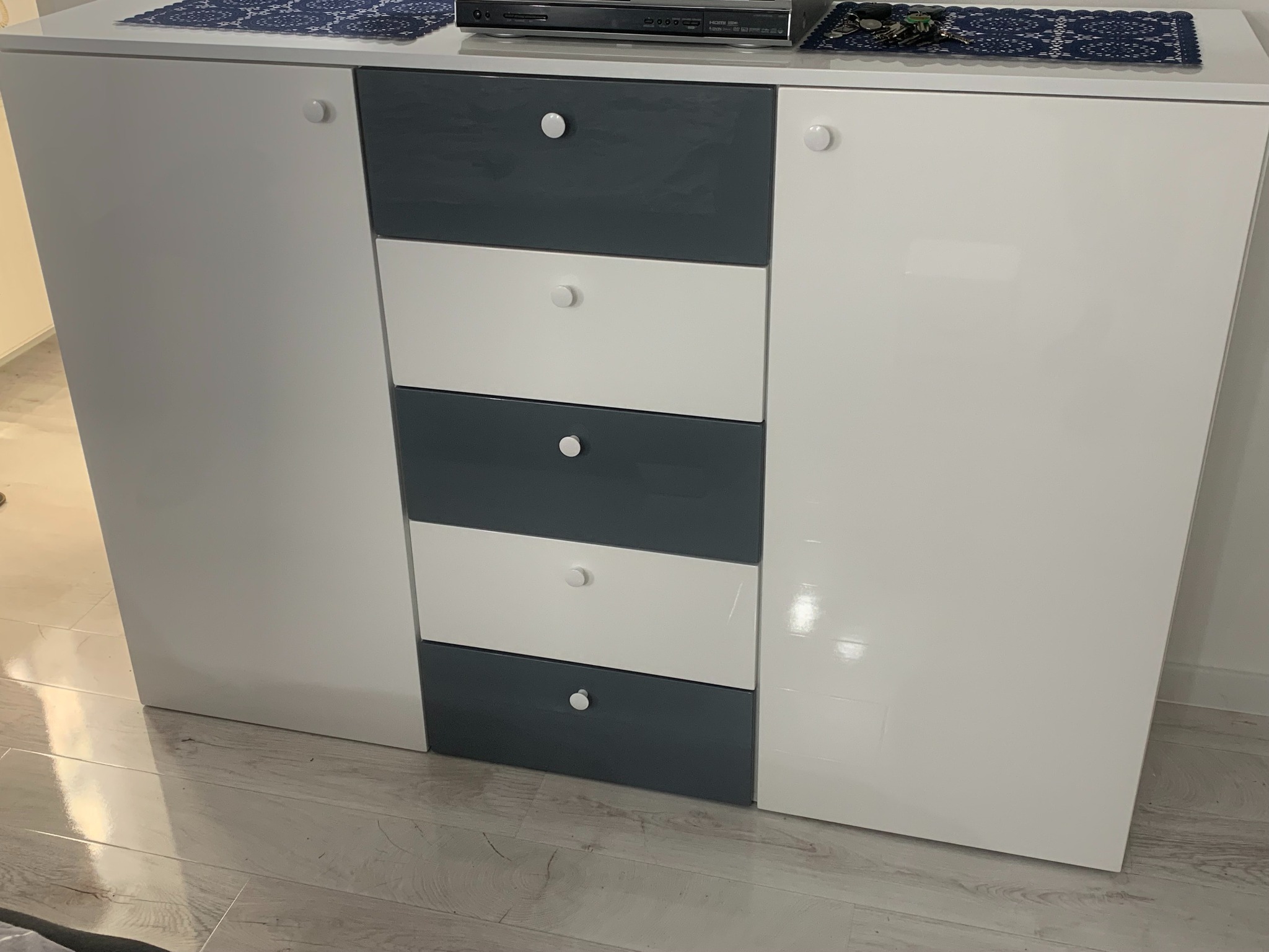 Nowoczesna komoda z szufladami i szafkami w kolorze białym i szarym, z okrągłymi uchwytami, zrealizowana w Gnieźnie. Na blacie widoczny odtwarzacz DVD i dekoracyjne serwetki.