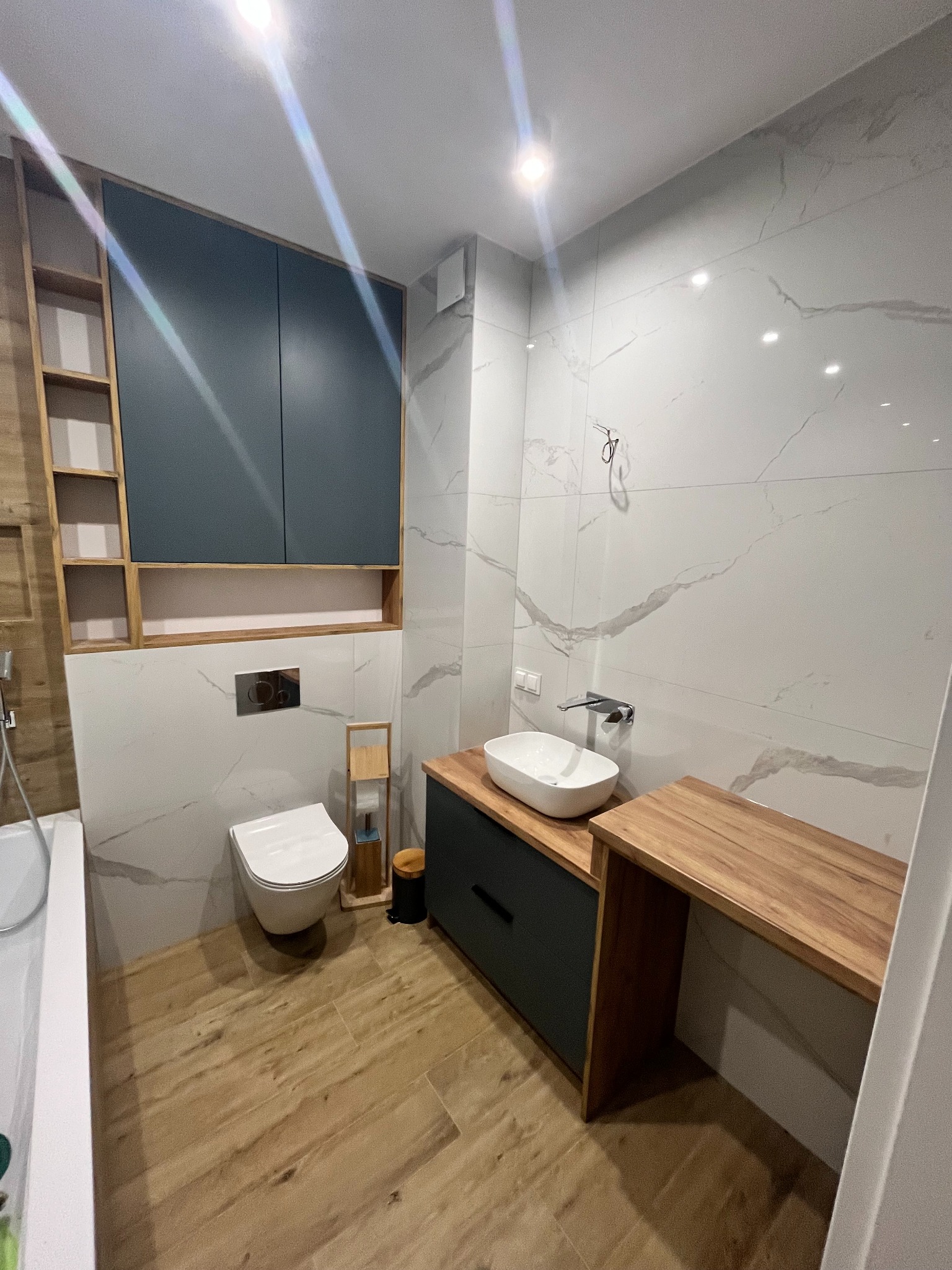 Nowoczesna łazienka z drewnianą podłogą, umywalką nablatową na szafce z szufladą, blatem z drewna i wiszącą szafką z ciemnymi frontami oraz otwartymi półkami z drewna.