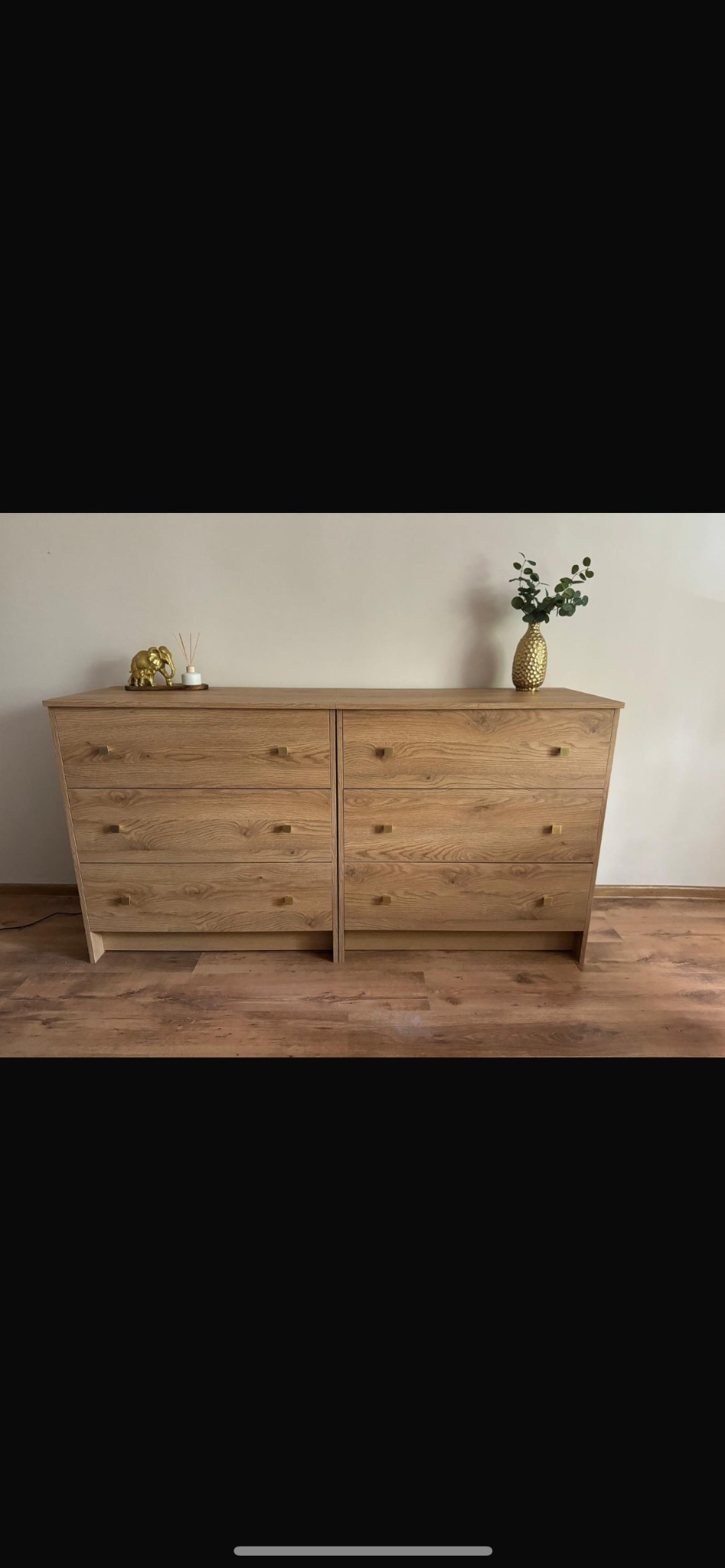 Komoda z jasnego drewna z sześcioma szufladami z kwadratowymi, złotymi uchwytami, na blacie złoty słoń i złoty wazon z zielonymi liśćmi, na podłodze panele.