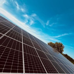 VARTECH RAFAŁ KASPRZAK - Czarno-szare panele słoneczne pod kątem, z widocznym błękitnym niebem z chmurami i zielenią drzew w tle.