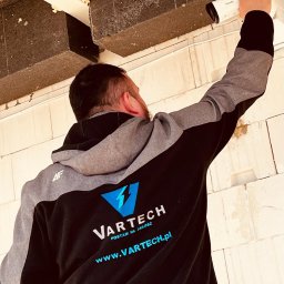 VARTECH RAFAŁ KASPRZAK - Instalator w bluzie firmowej montuje białą kamerę monitoringu na suficie z betonowych elementów konstrukcyjnych.