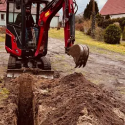 Czerwona mini koparka Yanmar Vio10-2A kopie wąski wykop w ziemi w Bytomiu Odrzańskim, widoczny operator w kabinie, w tle domy jednorodzinne i pochmurne niebo.