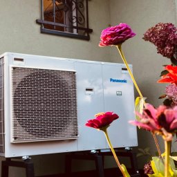 VARTECH RAFAŁ KASPRZAK - Biała zewnętrzna jednostka pompy ciepła Panasonic, umieszczona na czarnej metalowej podstawie, stoi na tle ściany budynku z małym oknem z ozdobną kratą; na pierwszym planie kolorowe kwiaty cynii...