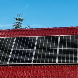 Panele słoneczne zamontowane na dachu z czerwonej dachówki, z widocznym oświetleniem zewnętrznym i pnącą roślinnością.