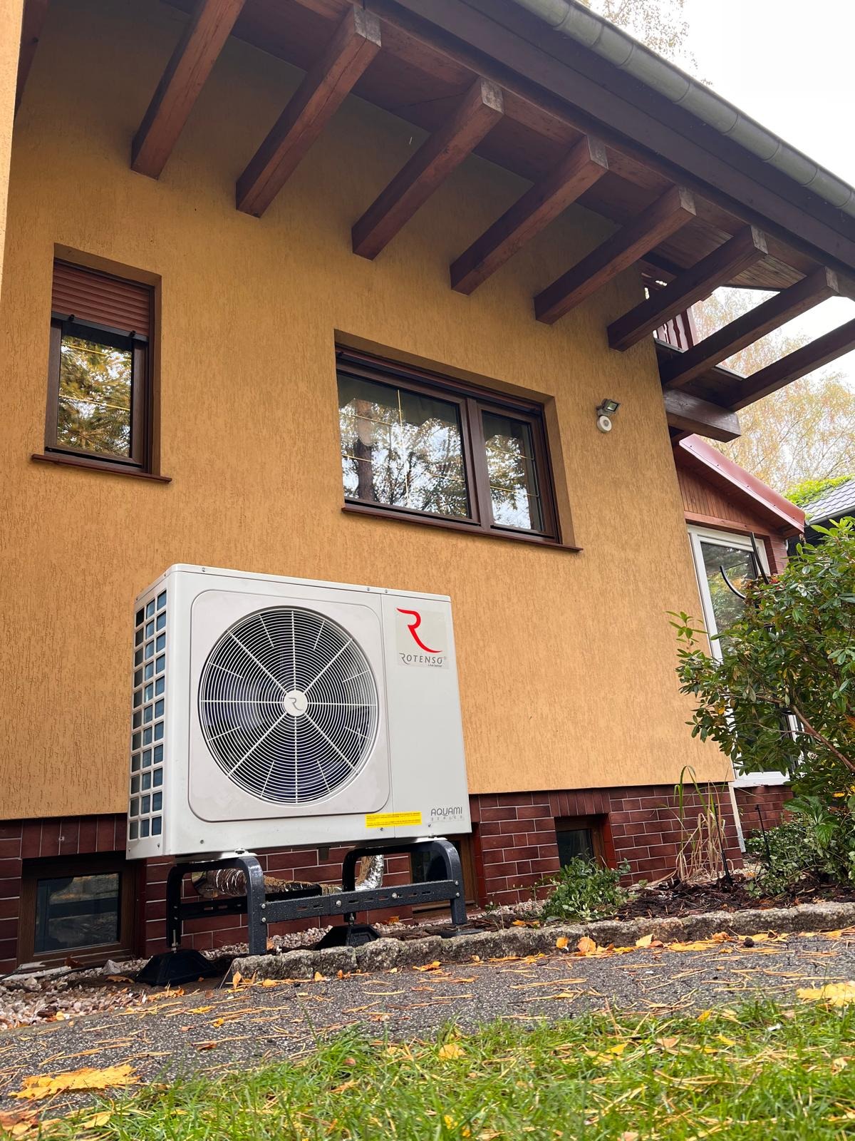 Biała pompa ciepła Rotenso Aquami zamontowana na zewnątrz budynku z beżową elewacją i brązowymi oknami, widoczna z poziomu trawnika pokrytego opadłymi liśćmi.