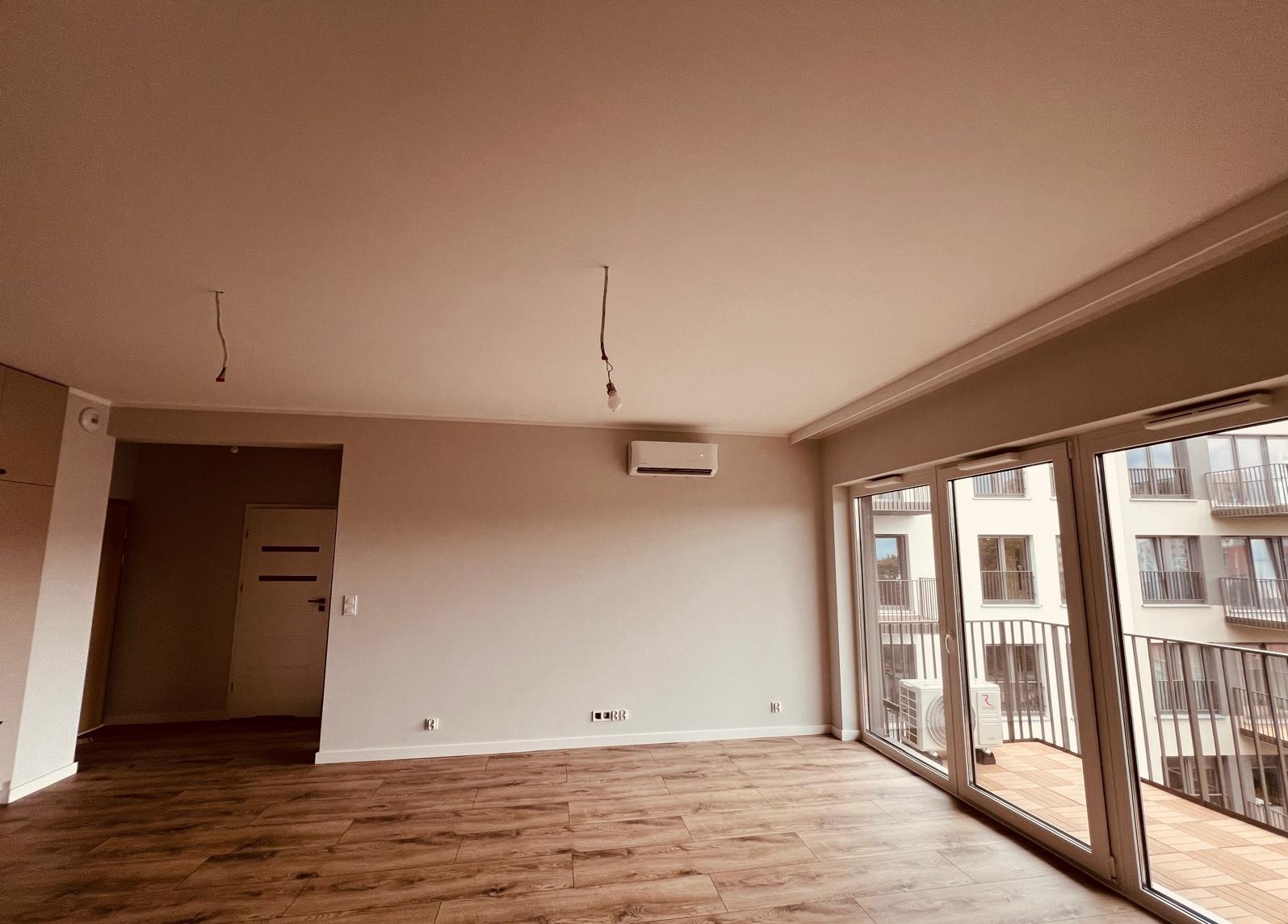 Nowoczesne wnętrze z zamontowaną klimatyzacją ścienną, widok na balkon z balustradą, drewnopodobna podłoga i surowe przewody elektryczne wiszące z sufitu.