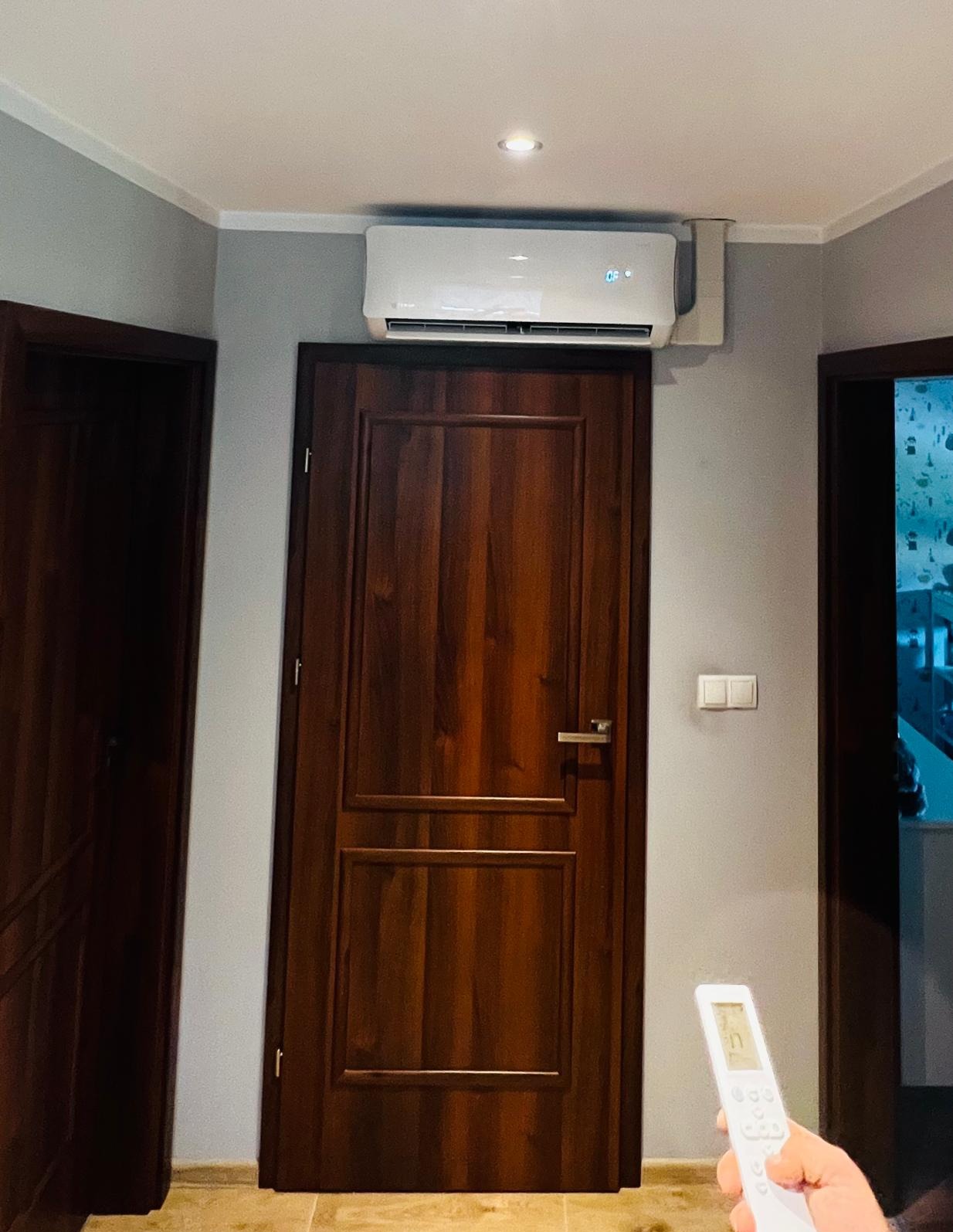 Biały klimatyzator zamontowany nad ciemnobrązowymi drzwiami w szarym pomieszczeniu, sterowany pilotem trzymanym w dłoni.