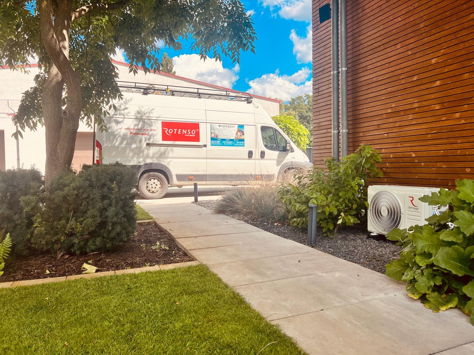Biały van firmy z logo Rotenso zaparkowany obok zainstalowanej jednostki zewnętrznej klimatyzatora przy budynku z drewnianą elewacją.