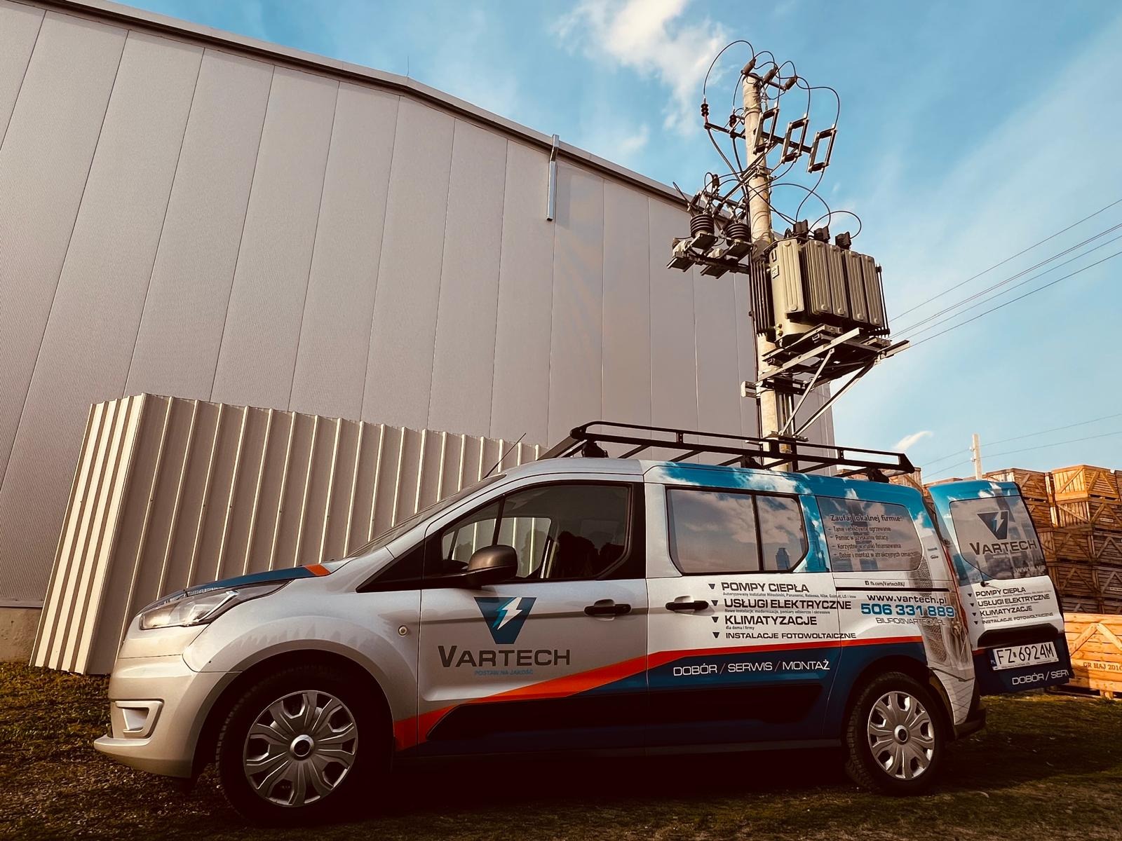 Srebrny samochód dostawczy z logo firmy VARTECH i reklamą usług: pompy ciepła, usługi elektryczne, klimatyzacja, instalacje fotowoltaiczne, zaparkowany przed przemysłowym budynkiem i słupem...
