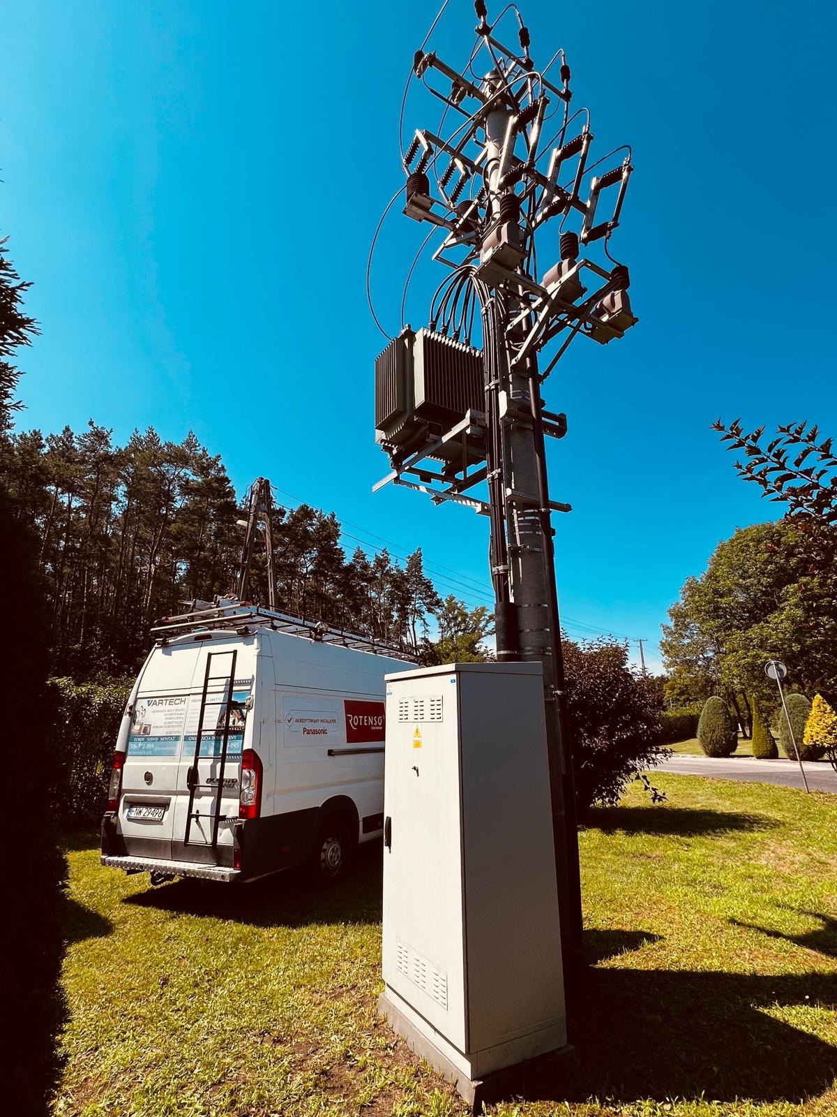 Słup energetyczny z transformatorem, skrzynka elektryczna i zaparkowany biały samochód dostawczy z drabiną na dachu na tle drzew i błękitnego nieba.