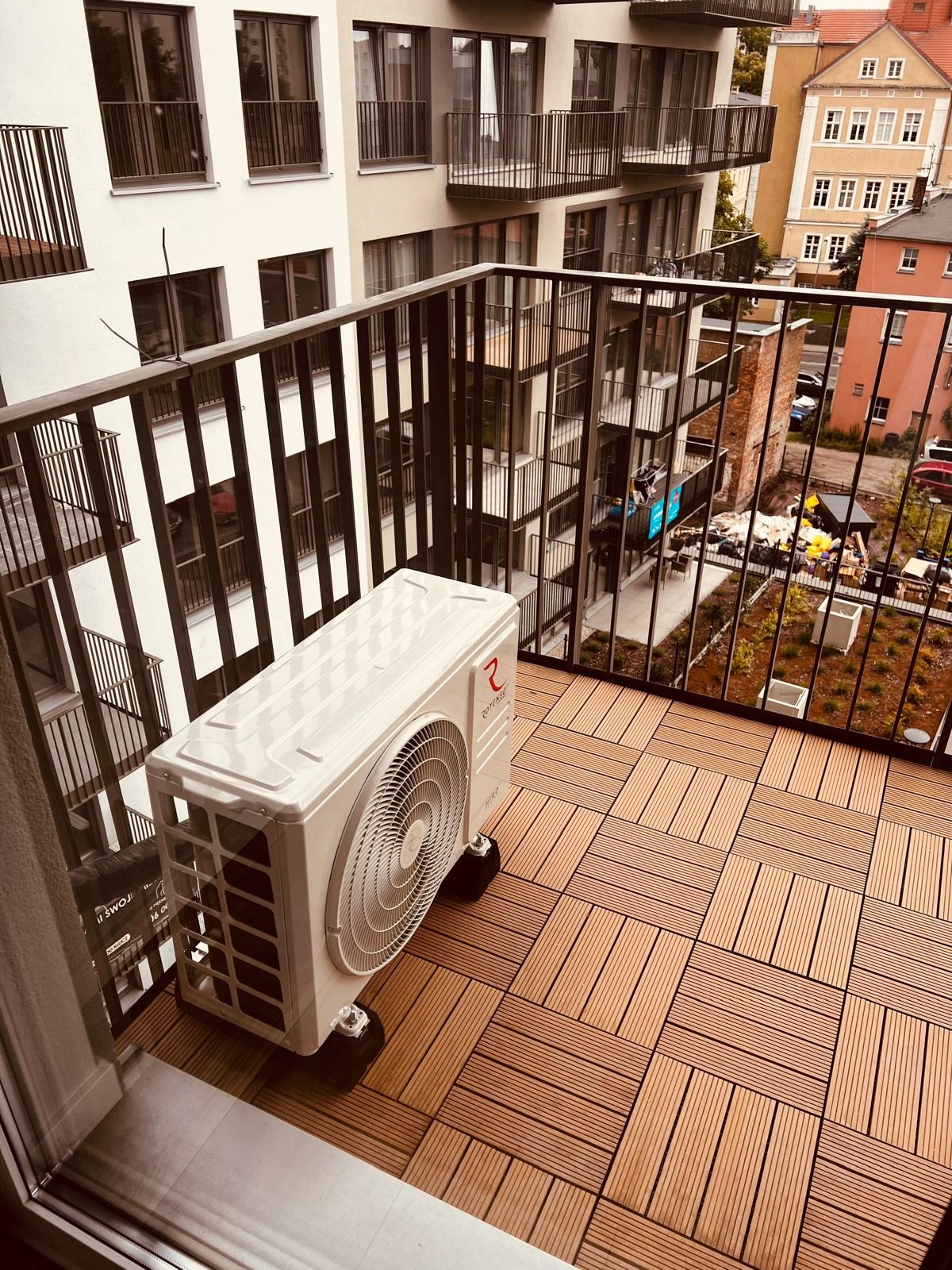 Biała jednostka zewnętrzna klimatyzatora na balkonie z brązową podłogą z płytek kompozytowych, widok na budynek mieszkalny i plac budowy.