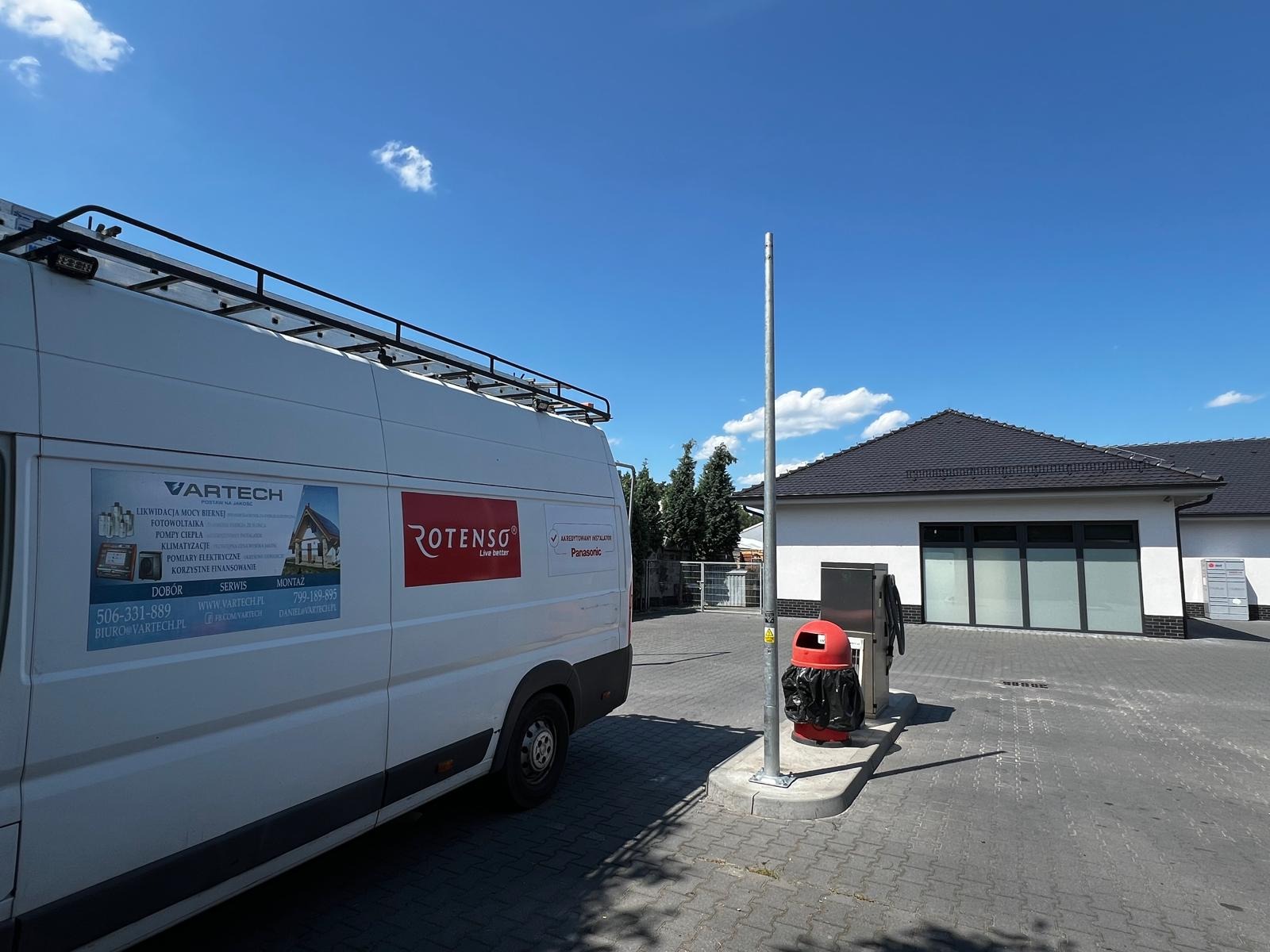 Biały samochód dostawczy z reklamą firmy Vartech oferującej fotowoltaikę, pompy ciepła i klimatyzację, zaparkowany na brukowanym parkingu przed budynkiem w słoneczny dzień.