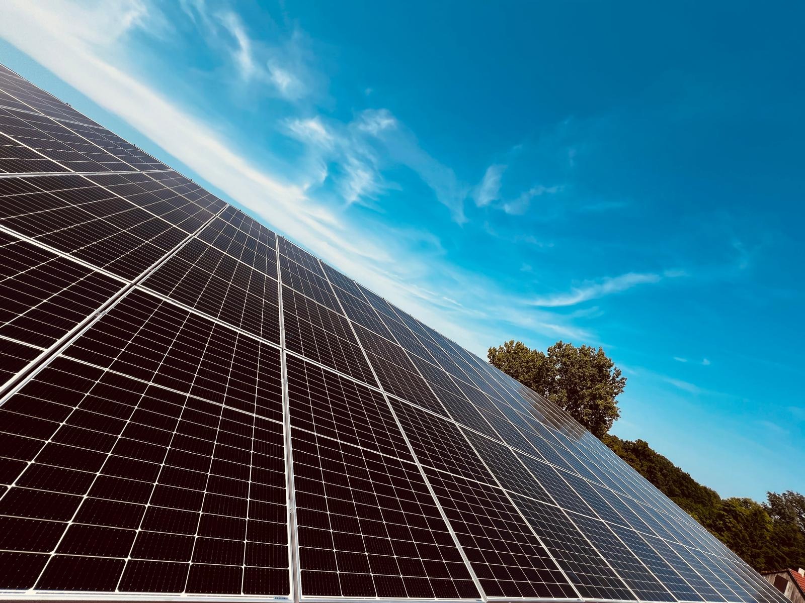 Czarno-szare panele słoneczne pod kątem, z widocznym błękitnym niebem z chmurami i zielenią drzew w tle.