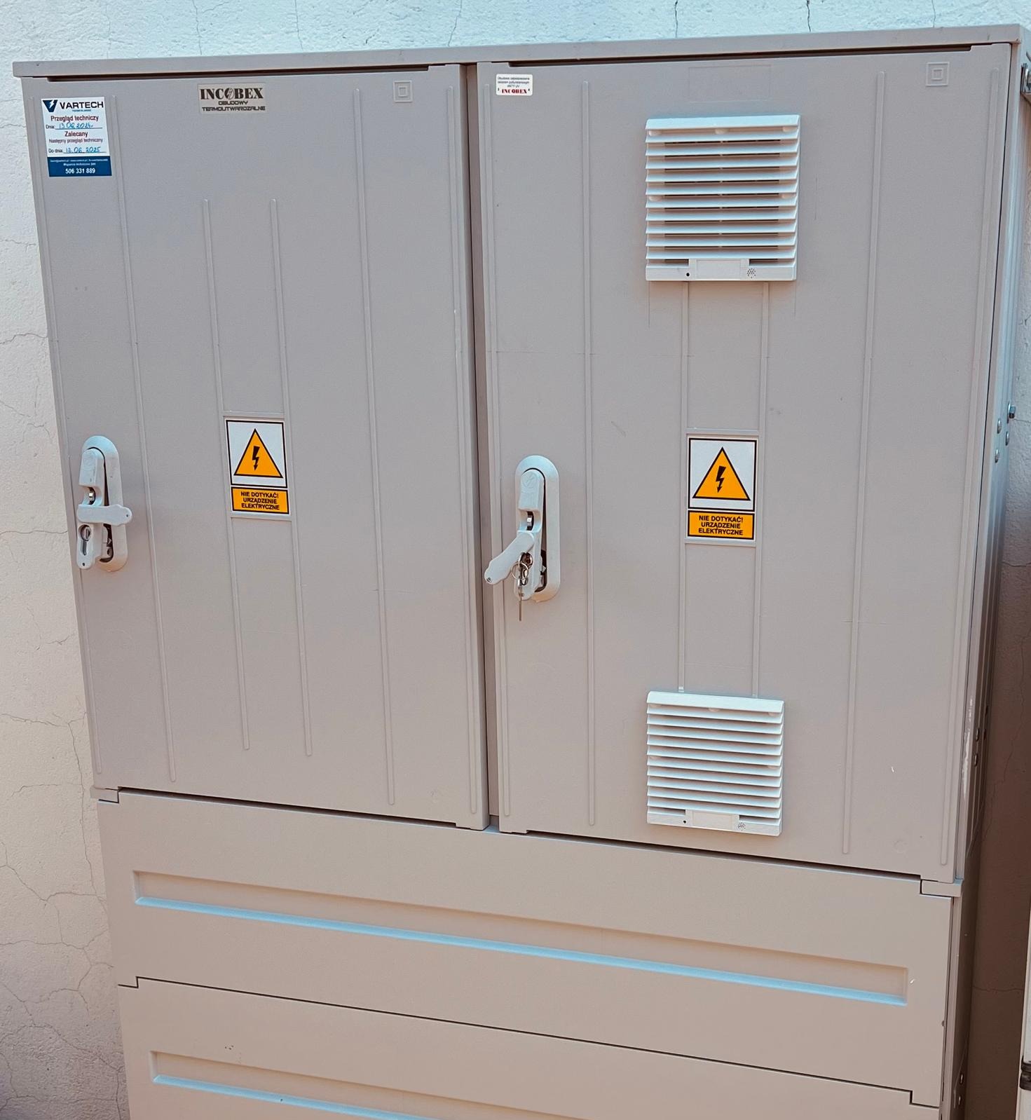 Szara szafa elektryczna z dwoma parami drzwi, wentylacją i ostrzeżeniami o wysokim napięciu, zamontowana na zewnątrz budynku.
