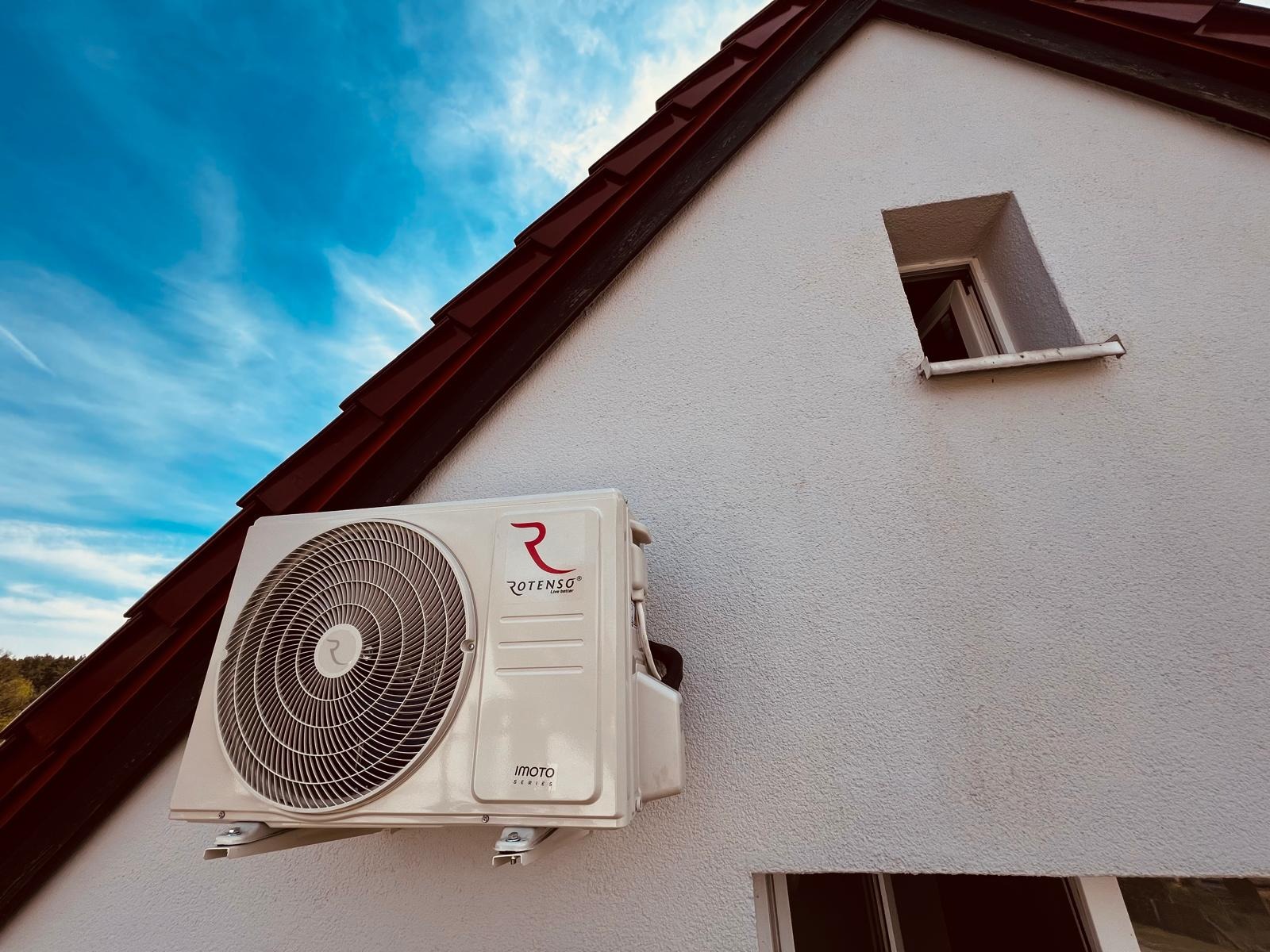 Zewnętrzna jednostka klimatyzacji Rotenso Imoto Series zamontowana na białej ścianie domu z czerwonym dachem, widoczna z dołu pod kątem, na tle błękitnego nieba.