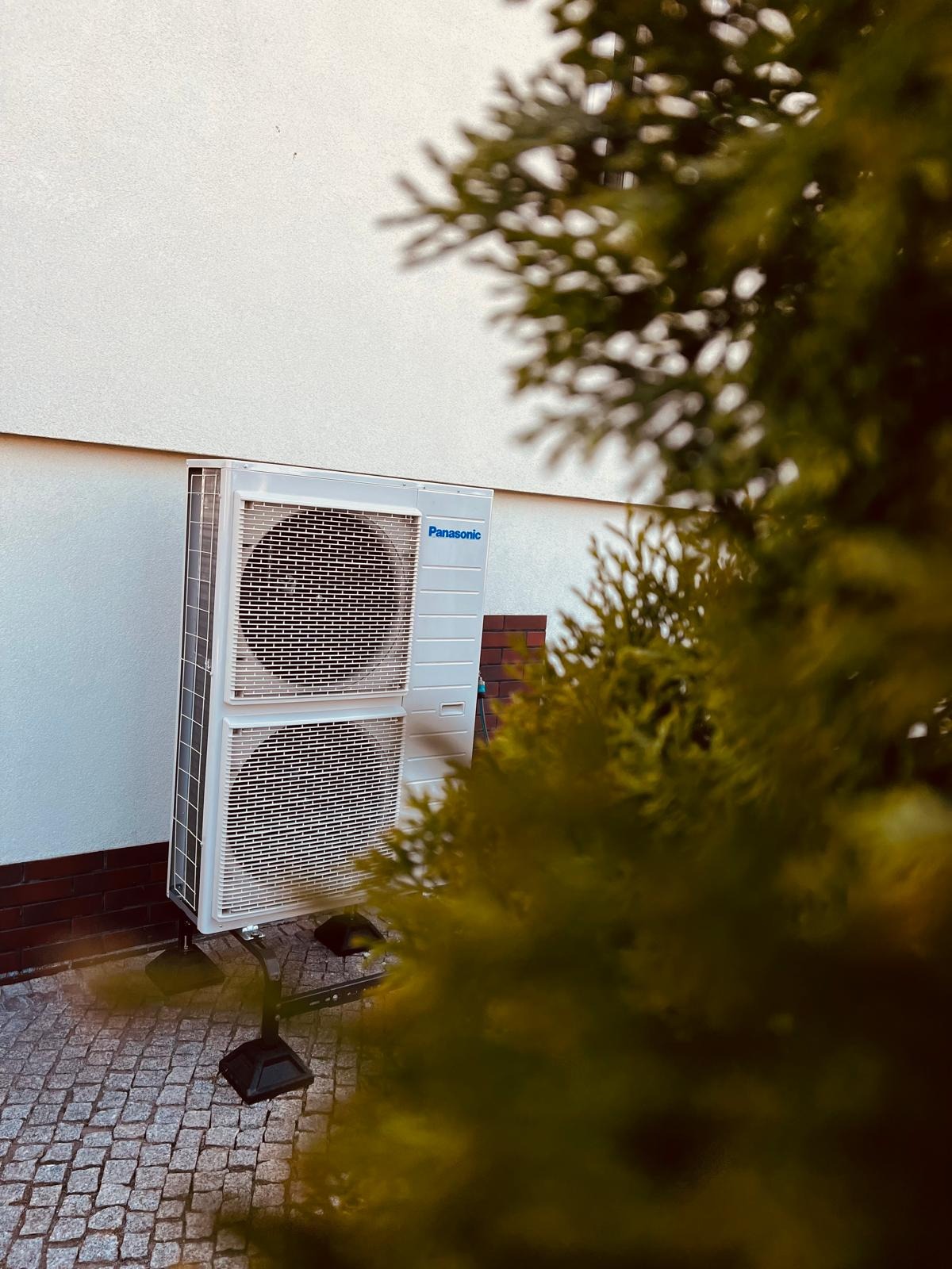 Biała jednostka zewnętrzna klimatyzacji Panasonic, zamontowana na wsporniku na kostce brukowej, widoczna przez gałęzie iglaka.