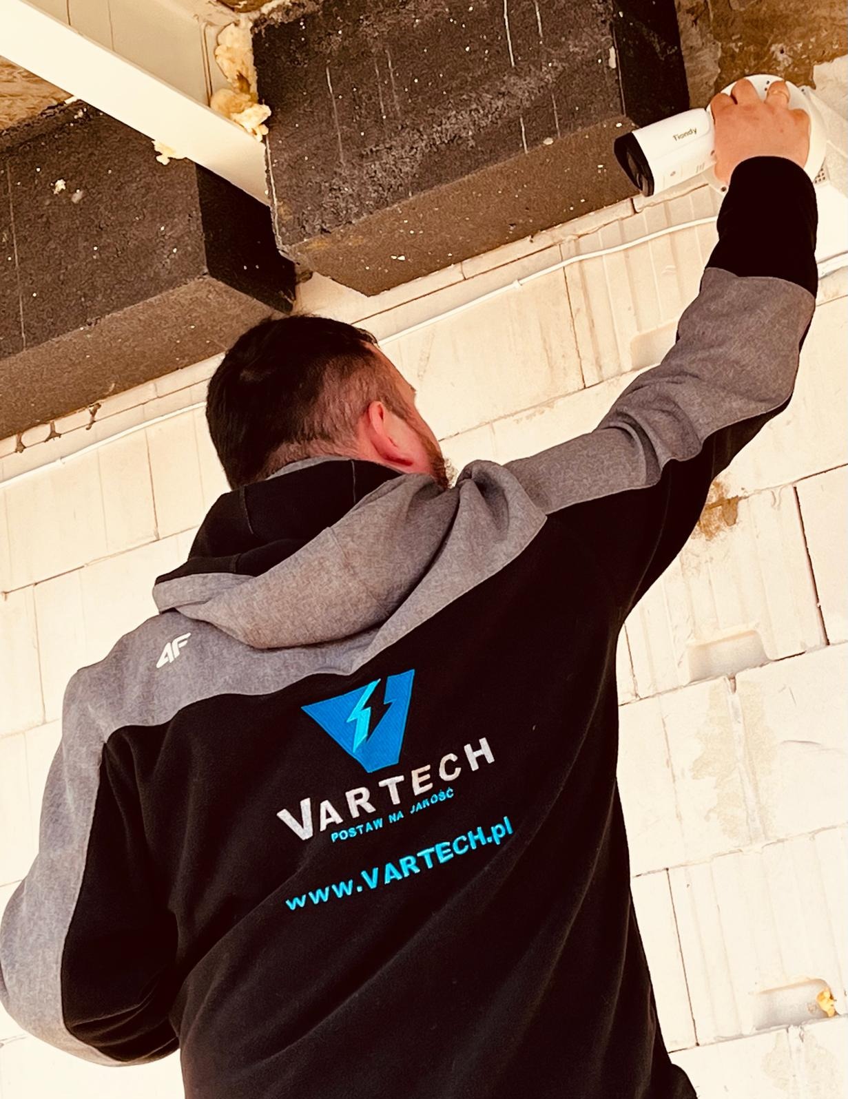 Instalator w bluzie firmowej montuje białą kamerę monitoringu na suficie z betonowych elementów konstrukcyjnych.