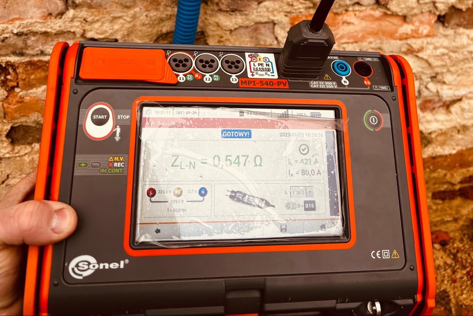 Miernik Sonel MPI-540-PV z wyświetlonym wynikiem pomiaru impedancji pętli zwarcia ZL-N = 0,547 Ω, wykonany na tle ceglanej ściany.