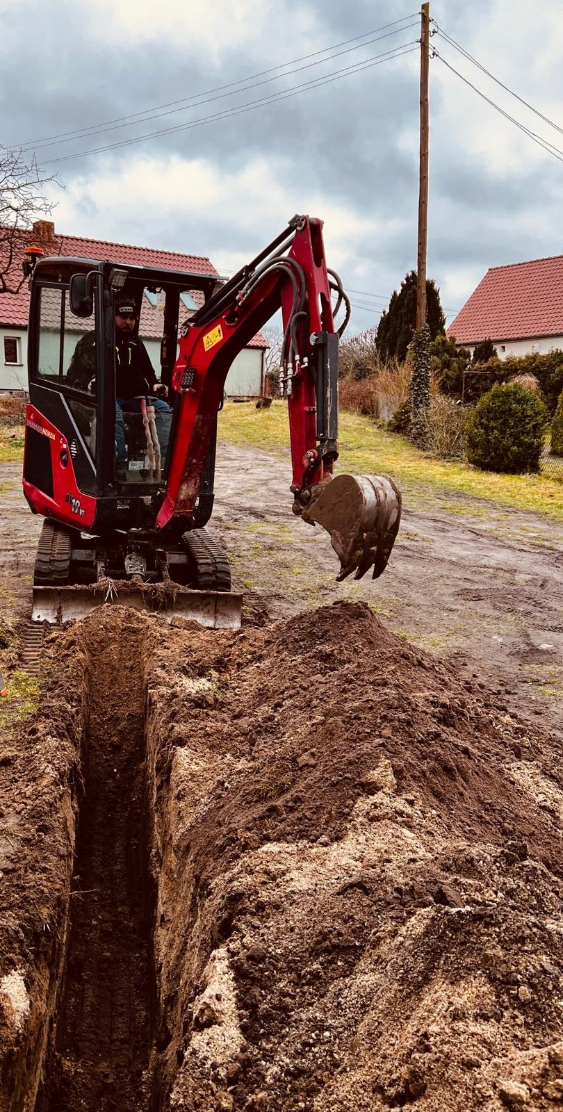 Czerwona mini koparka Yanmar Vio10-2A kopie wąski wykop w ziemi w Bytomiu Odrzańskim, widoczny operator w kabinie, w tle domy jednorodzinne i pochmurne niebo.