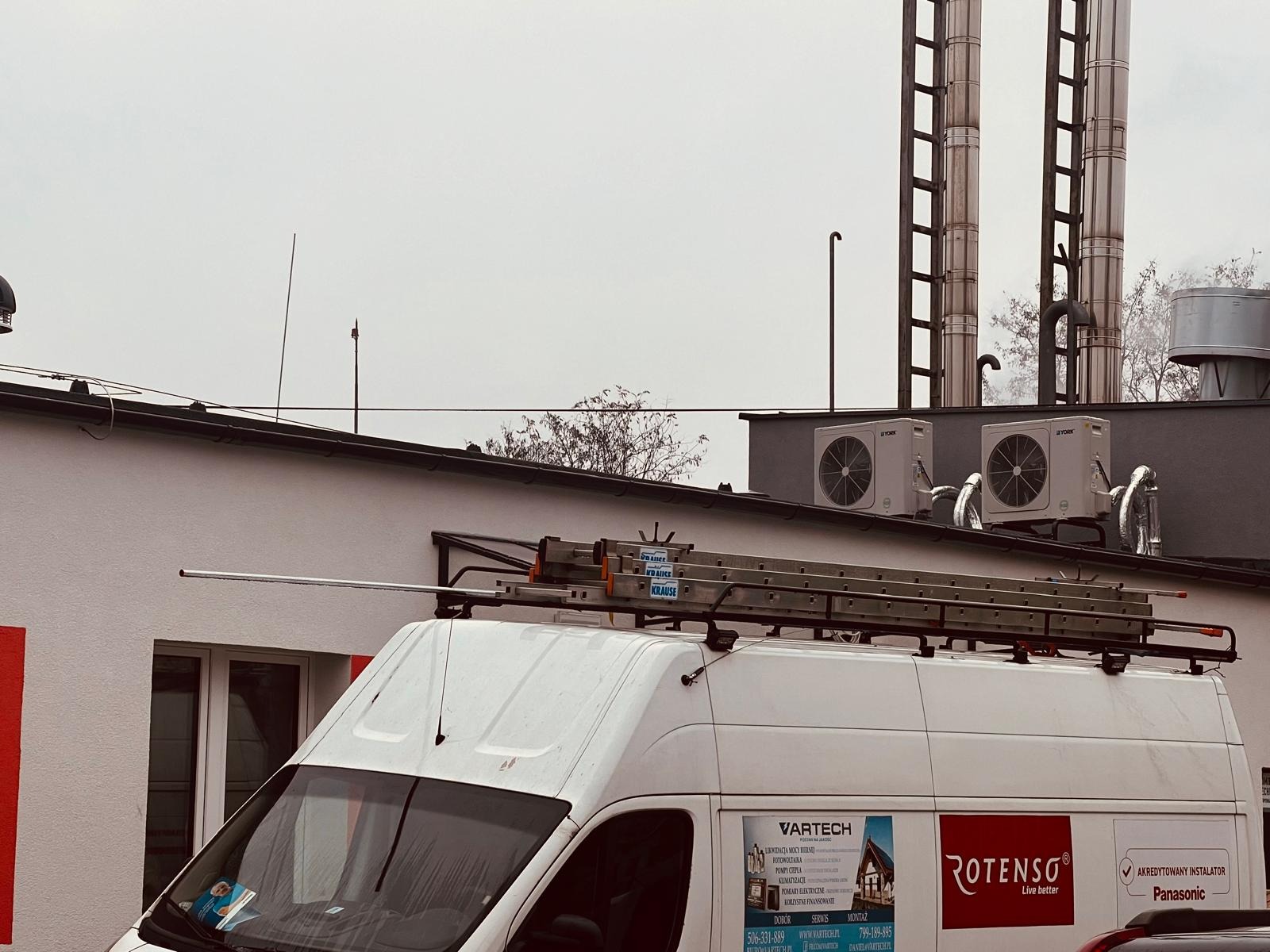 Biały samochód dostawczy z drabiną na dachu, na tle budynku z zamontowanymi jednostkami klimatyzacji i kominami wentylacyjnymi. Na samochodzie widoczne reklamy firmy instalacyjnej.