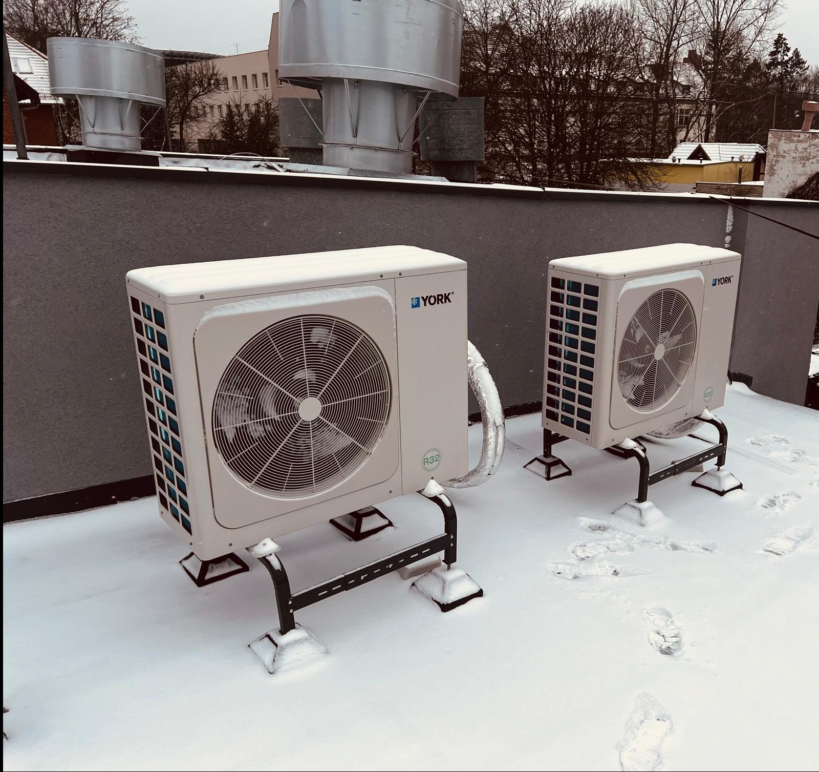 Dwa agregaty klimatyzacyjne marki York na dachu pokrytym śniegiem, widoczne ślady butów w śniegu, w tle budynki i drzewa.