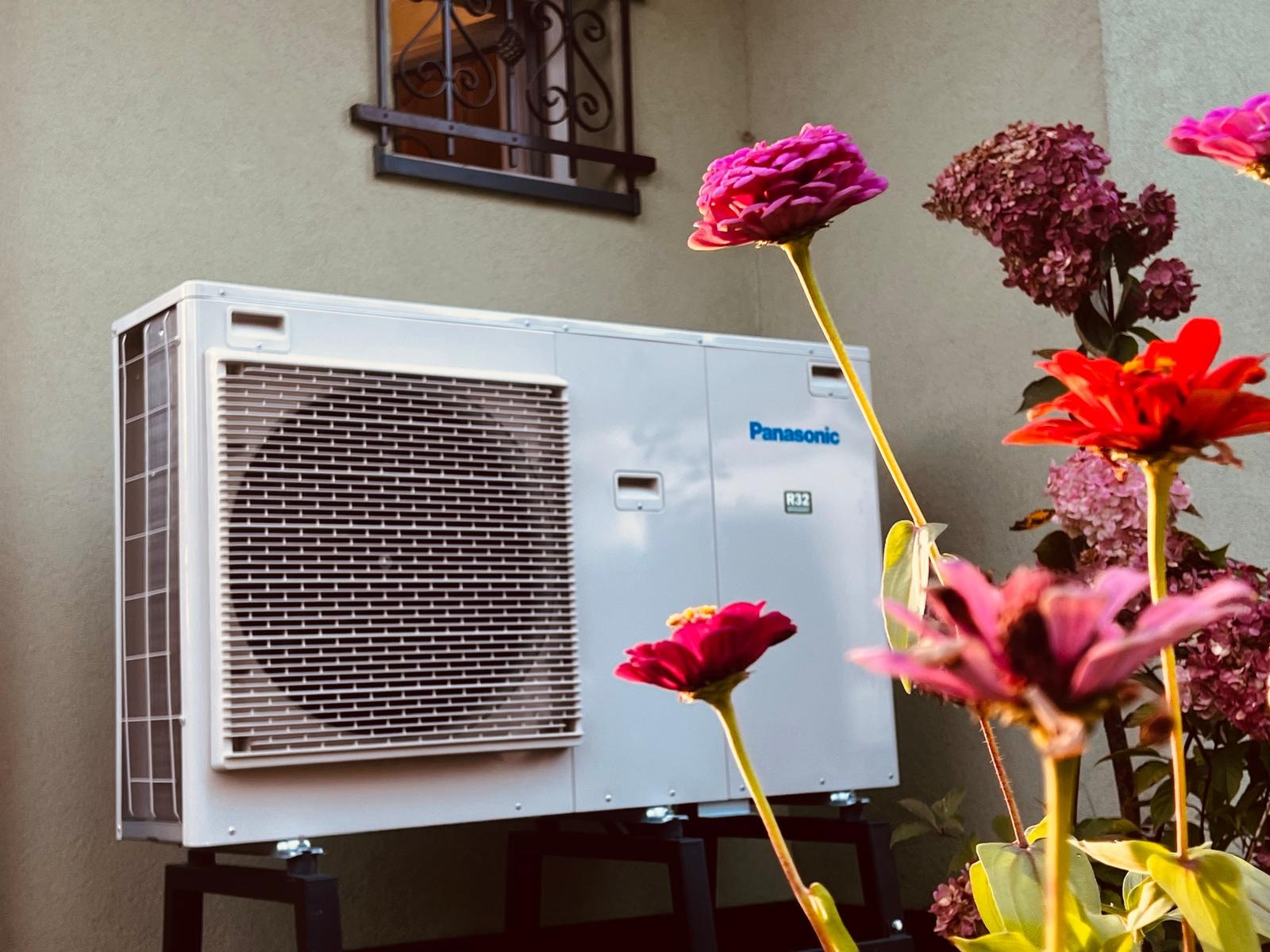 Biała zewnętrzna jednostka pompy ciepła Panasonic, umieszczona na czarnej metalowej podstawie, stoi na tle ściany budynku z małym oknem z ozdobną kratą; na pierwszym planie kolorowe kwiaty cynii...