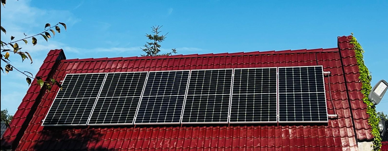 Panele słoneczne zamontowane na dachu z czerwonej dachówki, z widocznym oświetleniem zewnętrznym i pnącą roślinnością.