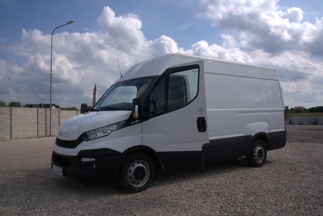 Biały samochód dostawczy marki Iveco na żwirowym parkingu z otwartymi drzwiami kierowcy, widoczne betonowe ogrodzenie w tle.