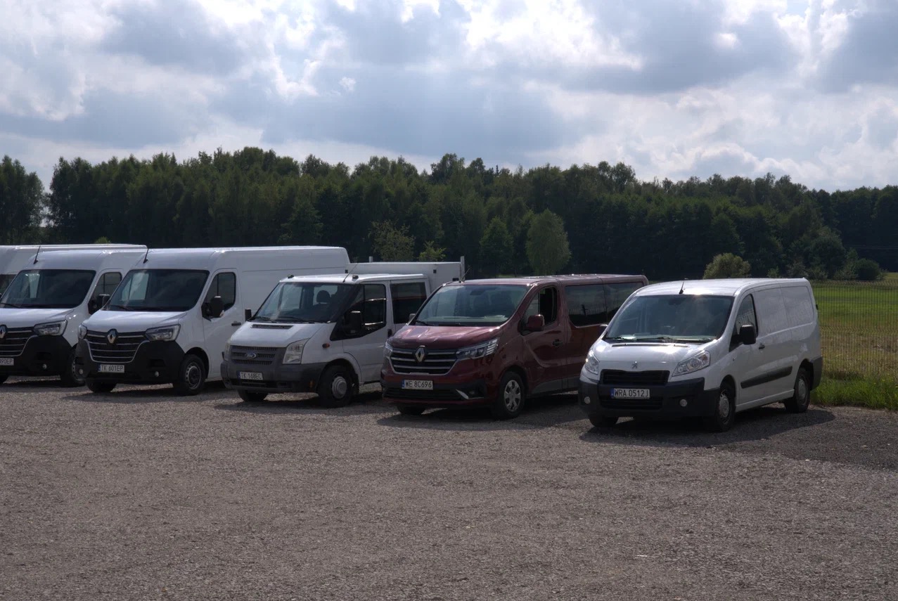 Rząd zaparkowanych samochodów dostawczych różnych marek i kolorów na żwirowym parkingu, z lasem w tle pod pochmurnym niebem.