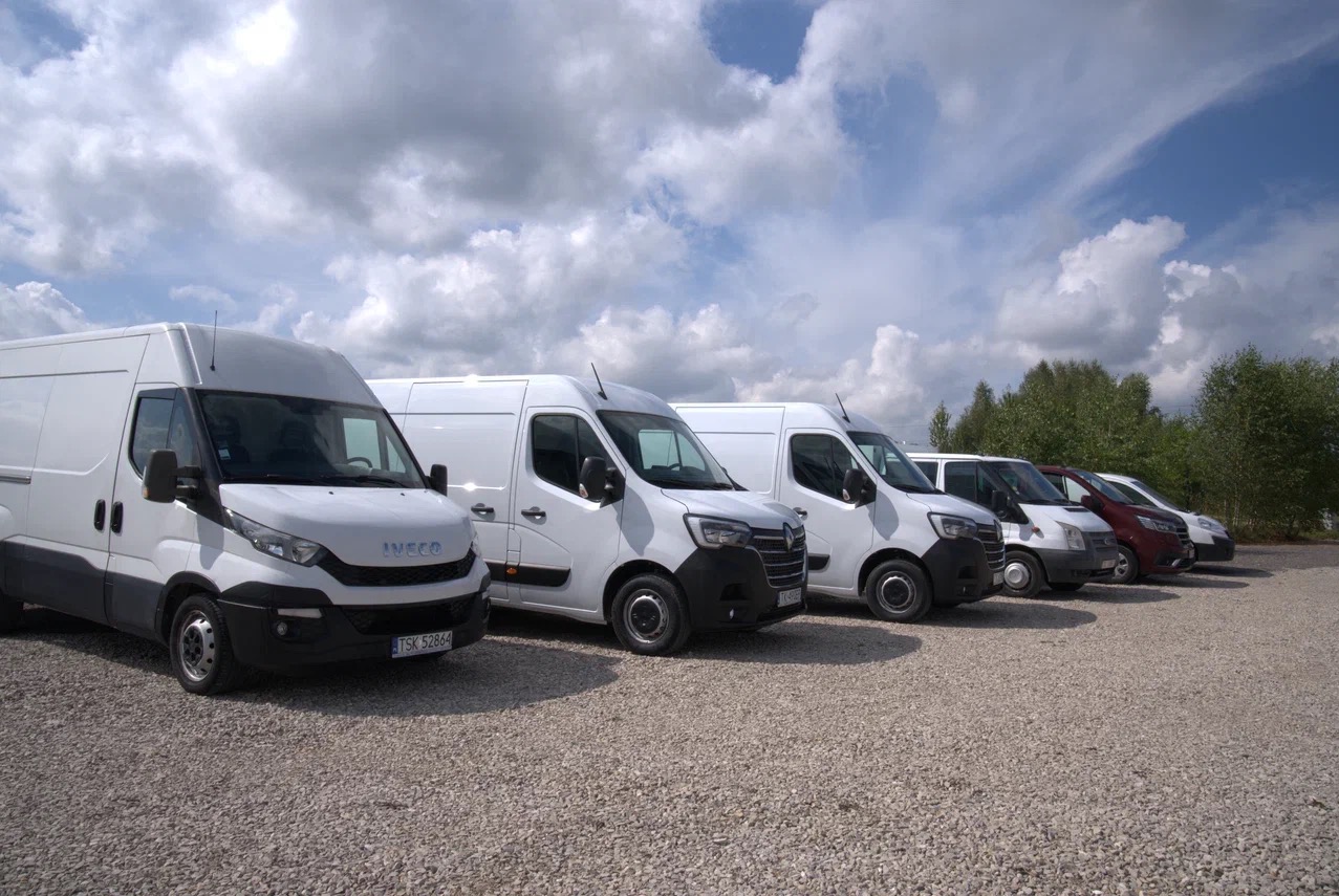 Rząd białych samochodów dostawczych różnych marek zaparkowanych na żwirowym parkingu w słoneczny dzień.