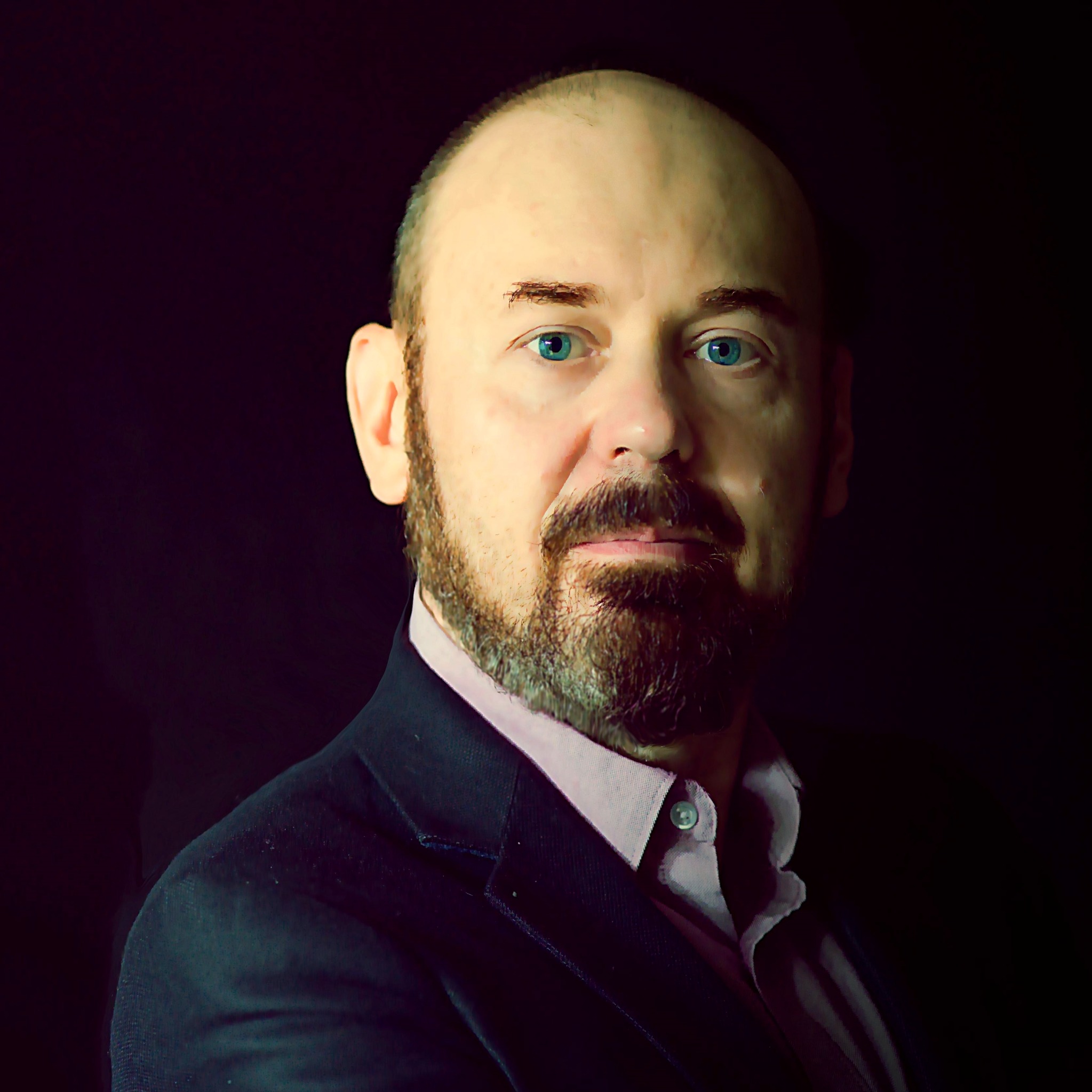 Portret mężczyzny z brodą i błękitnymi oczami, ubrany w ciemną marynarkę i jasną koszulę, na ciemnym tle.